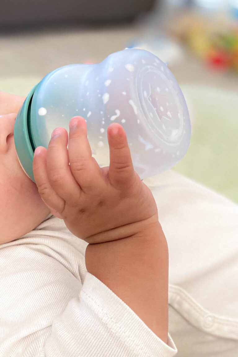 The image shows a close-up of a baby holding a Tommee Tippee bottle. The baby's hand is gripping the bottle, which is semi-transparent silicone with white milk droplets visible inside. The baby is dressed in a white onsie, and the background is softly blurred, focusing on the baby's hand and the bottle.