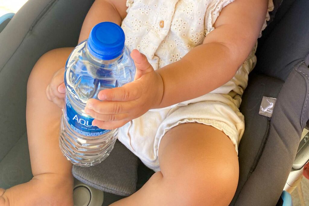 The photo shows a baby sitting in a black infant car seat, holding a standard Aquafina water bottle with both hands. The baby is dressed in a cream outfit with eyelet details. The water bottle is almost the size of the baby’s torso, making it appear oversized in the baby’s hands. 