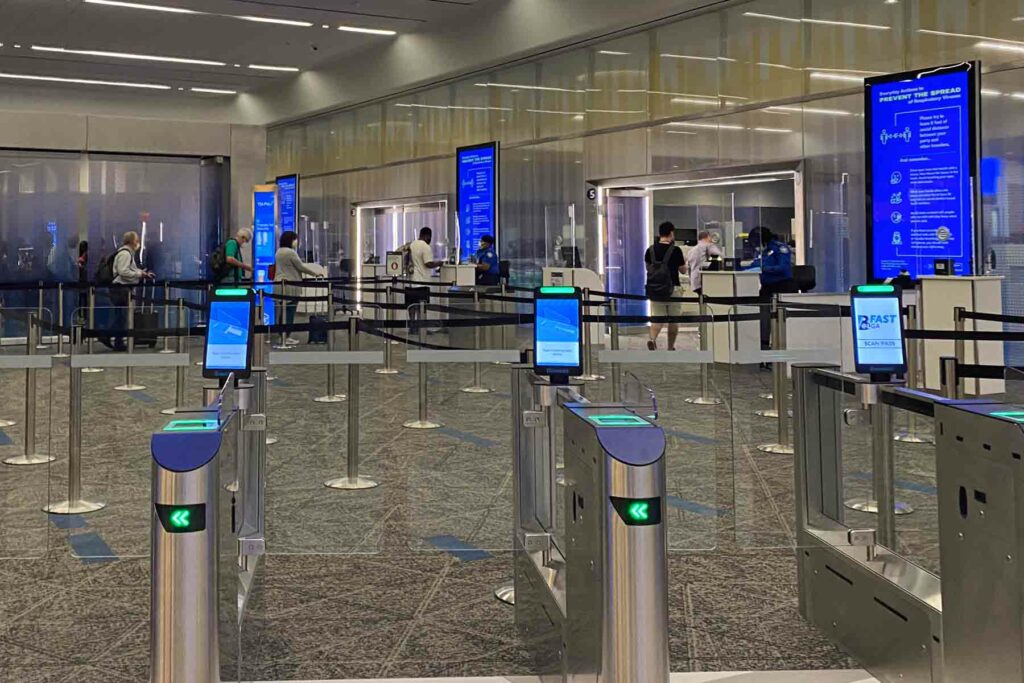 The image shows a modern airport security checkpoint with automated gates equipped with digital screens. The gates feature green arrow indicators and screens prompting travelers to scan their boarding passes. In the background, a few travelers are seen waiting in line, with large blue informational displays providing guidance on procedures. The airport looks very clean, modern and organized.