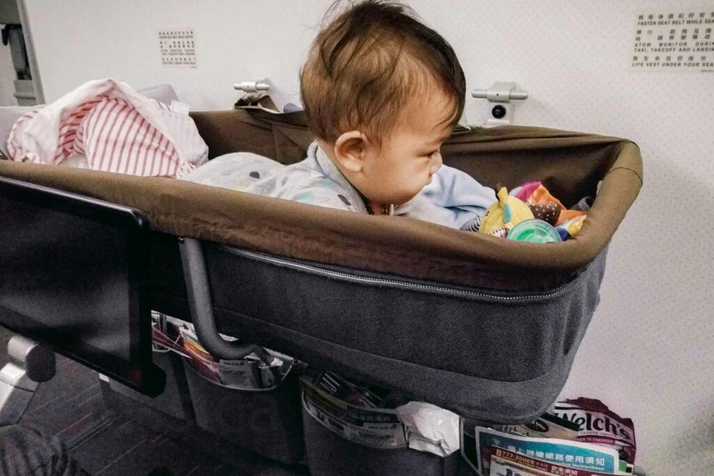 A baby lying on their tummy holding their head up in an airplane bassinet, playing with colorful toys. The bassinet is secured to the bulkhead, and various items, including a striped blanket, magazines, and snacks are stored in the plane pockets beneath it.