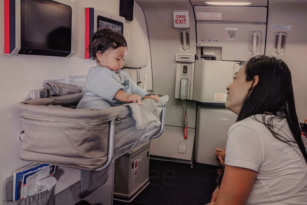 A 6 month old baby sits in an airplane bassinet on a long haul flight while their mom sits across the seat, smiling. The baby, dressed in a light blue sleepsuit, plays with the edge of the bassinet. Behind them, the airplane’s emergency exit and various cabin features are visible.