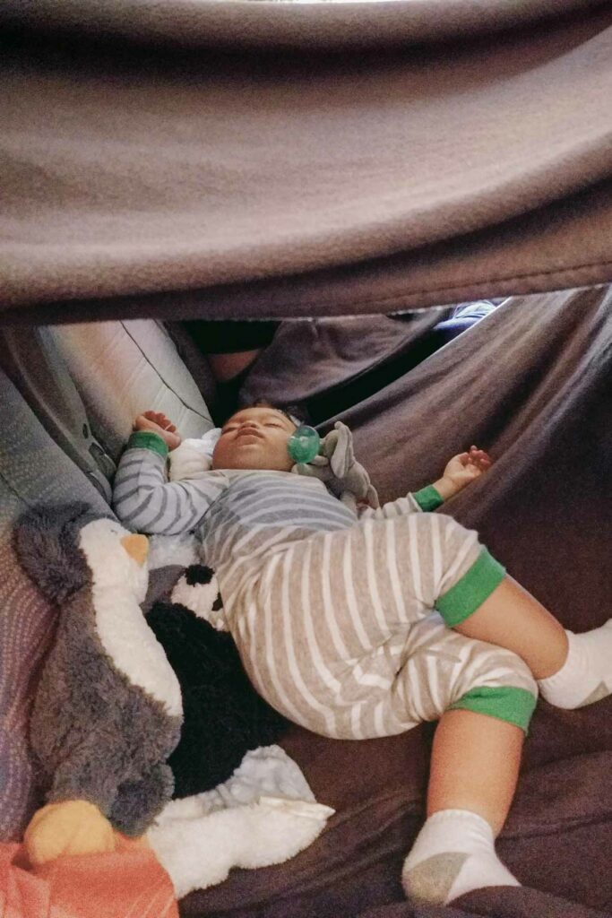 A baby wearing white and gray striped pajamas with green cuffs sleeps peacefully on a long-haul flight inside a makeshift tent created by several blankets. The baby is surrounded by plush stuffed animals, including a penguin and panda, and has a pacifier nearby.