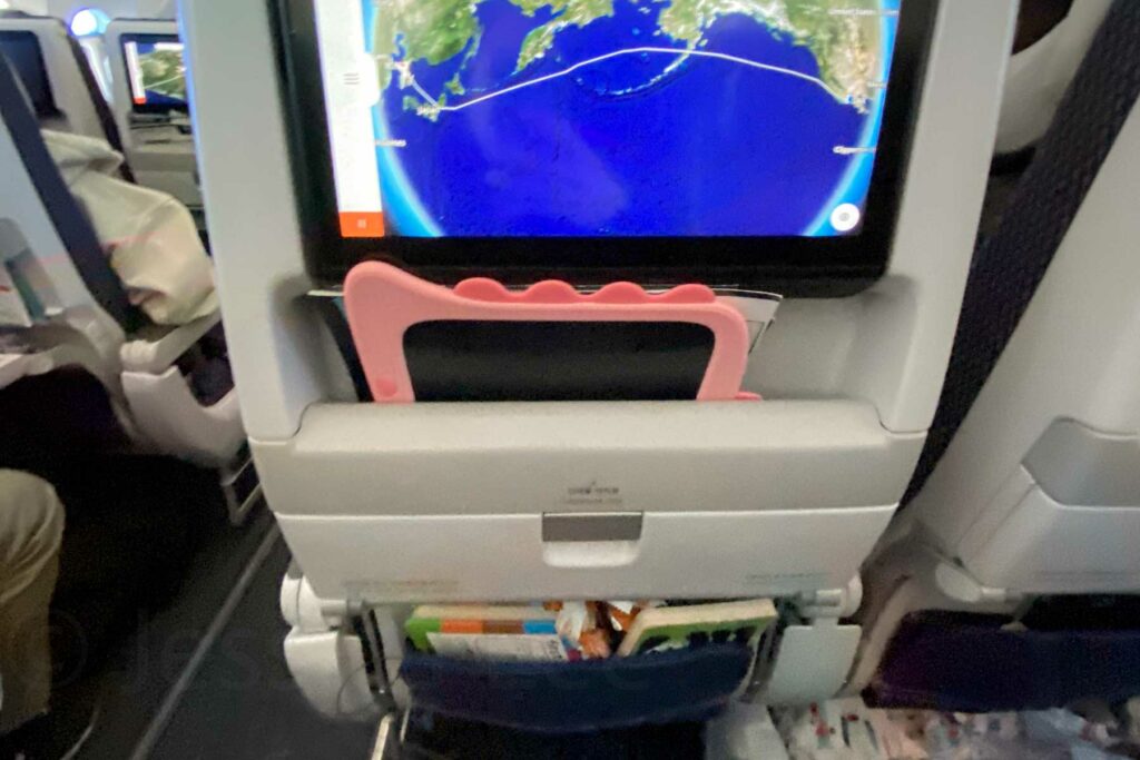 View of the back of an airplane seat showing an in-flight screen displaying a world map with a flight route crossing over the Pacific Ocean. A pink drawing tablet is tucked in the upper seat pocket while the lower seat pocket has more items, including snacks, books, and brochures.