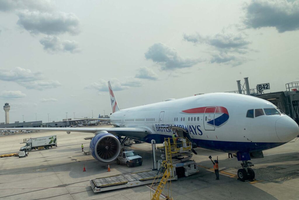 A British Airways airplane is parked at the gate, with a jet bridge and loading vehicles surround it. The sky is overcast with scattered clouds, and an airport control tower is visible in the distance.