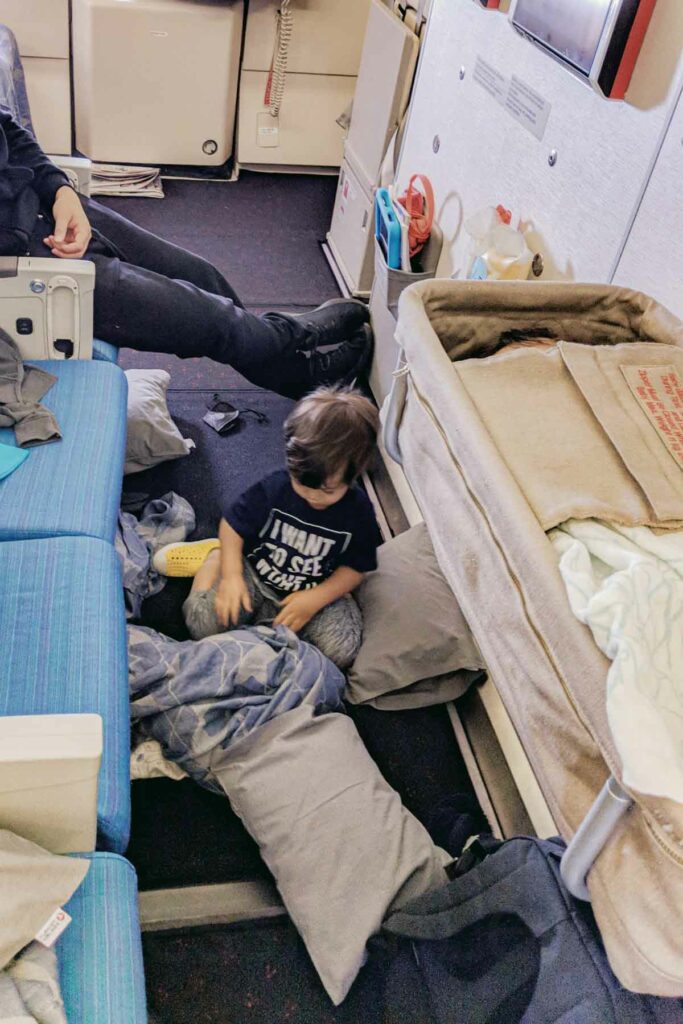 A toddler on a long haul flight wearing a navy graphic tee, gray sweatpants, and yellow shoes plays on the floor of the bulkhead seat area on an airplane, surrounded by pillows and blankets. To the right, a baby is asleep in an airplane bassinet attached to the wall. An adult sits in the nearby seat, with their feet propped up, watching over the children.