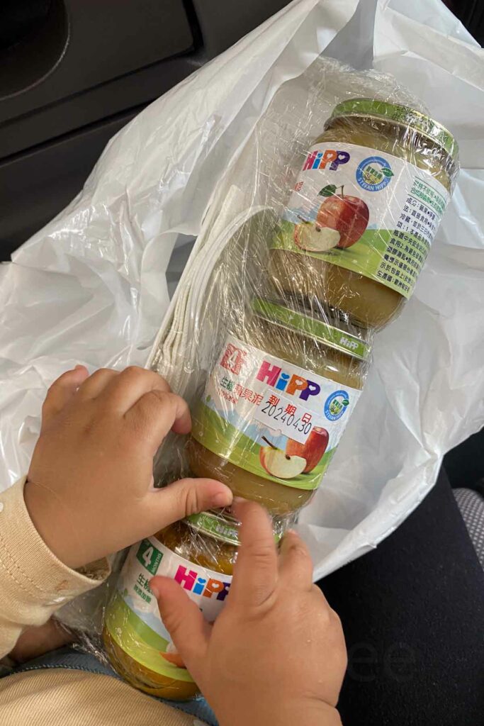 Close-up of a baby’s hands holding a plastic-wrapped bundle of HiPP baby food jars while sitting on a plane. The jars are labeled with an apple flavor, with the packaging displaying product information in Chinese.