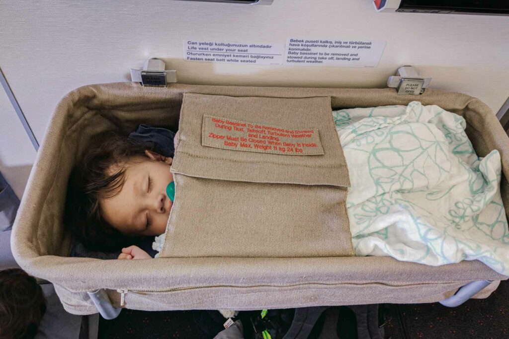 A baby is sleeping peacefully inside an airplane bassinet on a long-haul flight, with a green pacifier in their mouth. The bassinet has a label stating weight limits and safety instructions, and a light muslin with and blue blanket covers part of the baby.