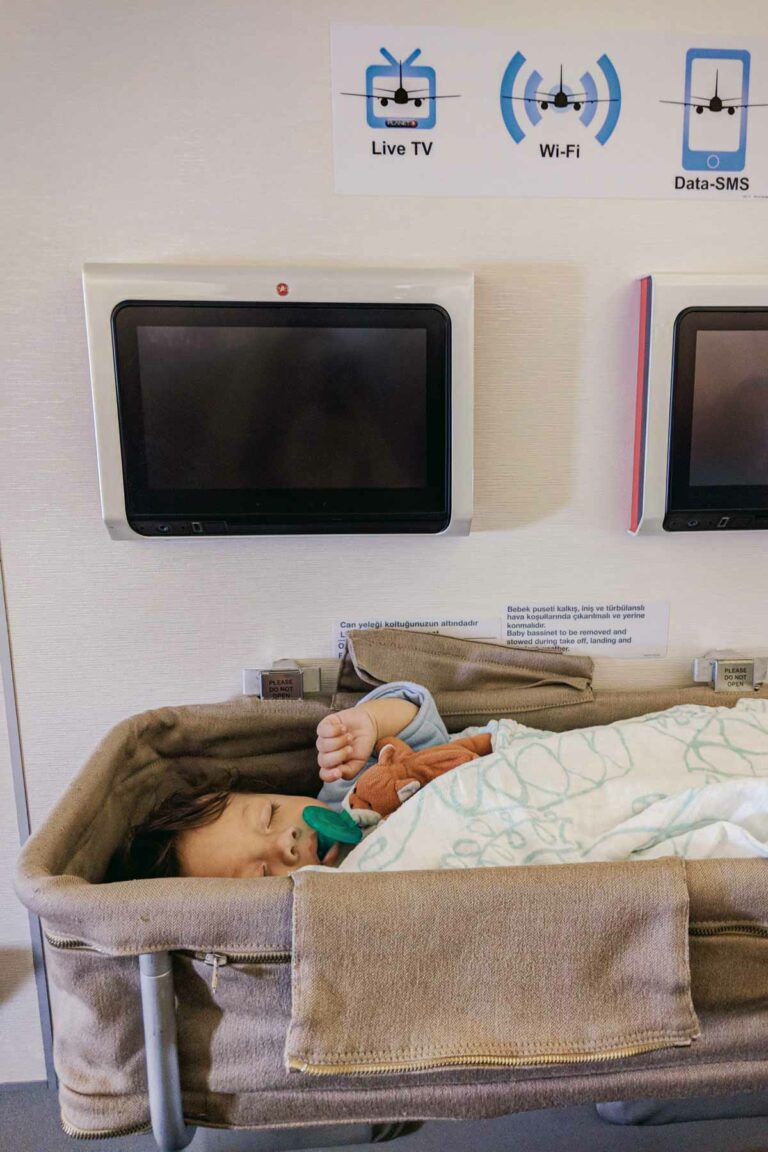 A baby is sound asleep in an airplane bassinet on a long flight on a Turkish Airlines plane, with a pacifier in their mouth and wrapped in a light blanket. Above the bassinet, two in-flight entertainment screens are mounted on the wall, along with signs for Live TV, Wi-Fi, and Data-SMS.