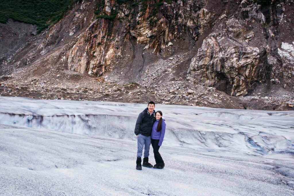 Couple standing on the Mendenhall Glacier in Alaska, with dark rocky cliffs in the background which contrasts with the bright white and slightly blue glacier under their feet. Both are dressed in winter gear, smiling and posing closely together on the icy surface.