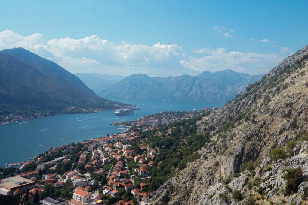 Scenic view of the Bay of Kotor in Montenegro, with towering mountains, a cruise ship docked, the red roofs of buildings, and rocky mountains set against a bright blue summer sky.