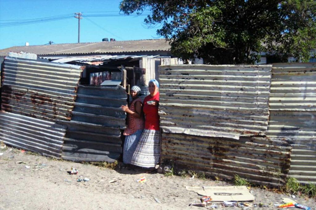 Two South African women standing in the opening of a corrugated metal wall fence in Cape Town, South Africa, looking at the camera with their skirts blowing in the wind.