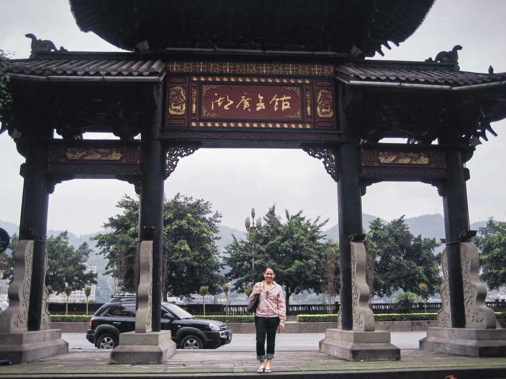 Female traveler in a light pink coat and jeans standing in front of a traditional Chinese gate in Chongqing, China. The ornate gate has intricate carvings and inscriptions, with trees and a parked car visible in the background.