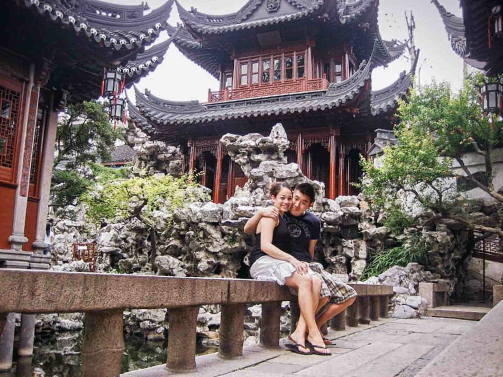 Couple sitting on a stone bench in Yuyuan Garden, Shanghai, China. The traditional Chinese architecture features red paint, black ornate roofs, and intricate wooden details, surrounded by lush greenery and rock formations.