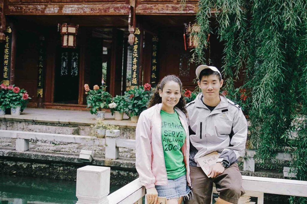 Young couple posing on a bridge in Yunnan, China. Behind them is a traditional wooden building adorned with potted plants and Chinese inscriptions, with a weeping willow tree hanging over the jade green water.