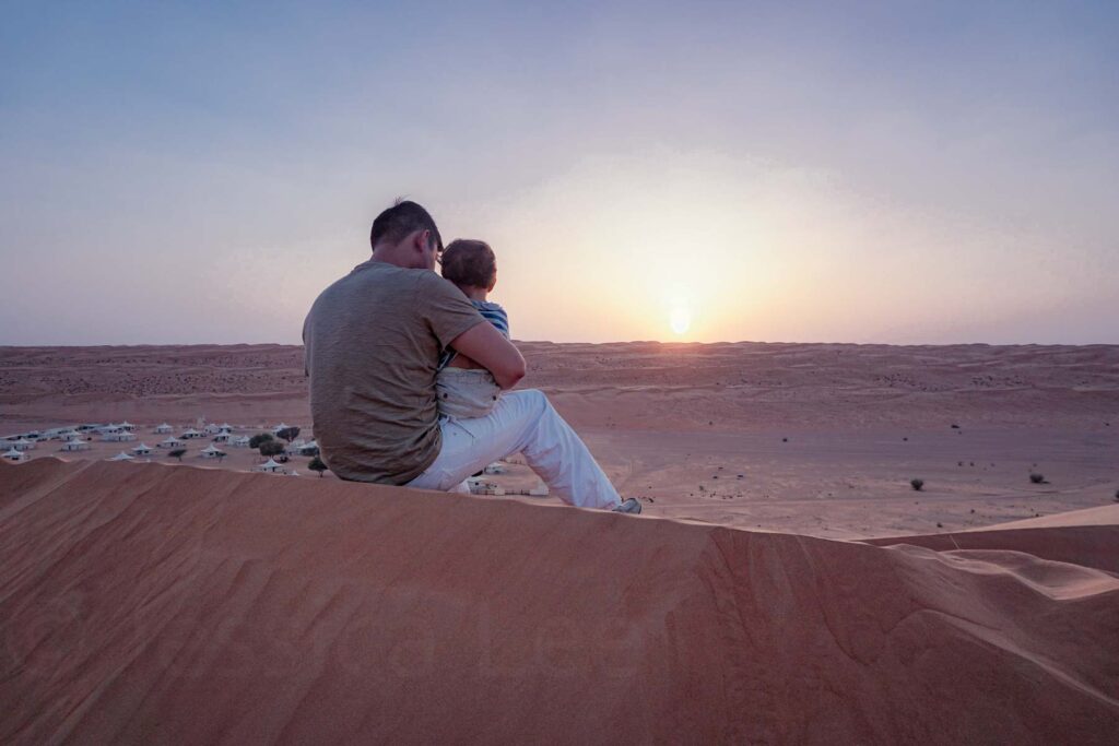 A father holding his young son on his knee perched above a desert camp watching the sunset over the vast sand dunes of the Wahiba Sharqiya Sands in Oman.