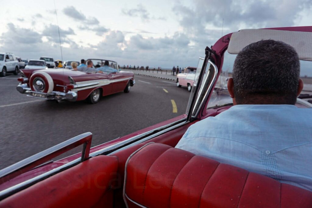Classic American red Chevy convertibles driving along the Malecon in Havana, Cuba, with the sea and cloudy sky in the background at dusk