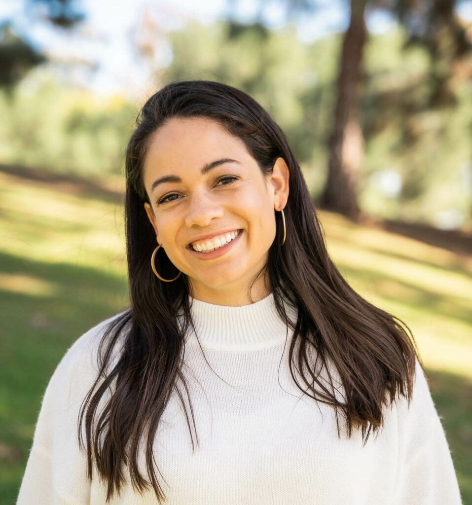 Mixed woman with long straight dark brown hair looking straight on the camera smiling wearing a white sweater with a trees a park blurred in the background