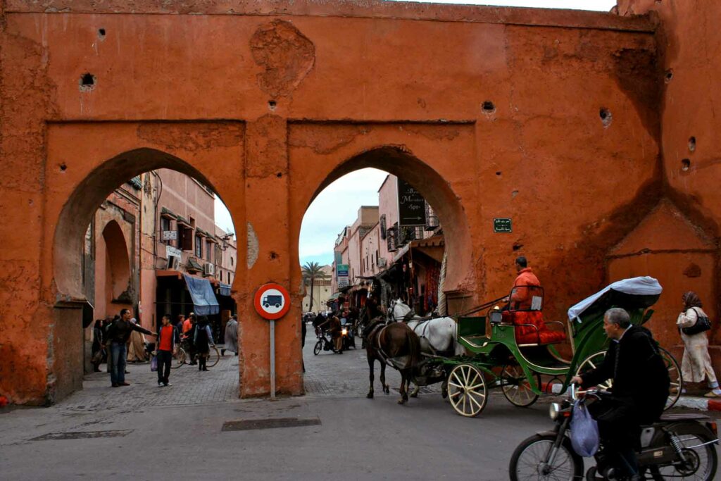A street scene in Marrakesh, Morocco, with horse-drawn carriages and people passing under historic archways of the red walls.