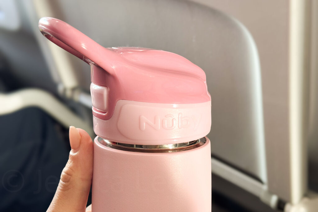 The image shows a close-up of a pink Nuby stainless steel toddler bottle being held by a person, with the cap prominently featuring a flip-up straw. The cap is slightly unscrewed. The background shows the interior of an airplane, with an out-of-focus airplane seat and tray table visible.