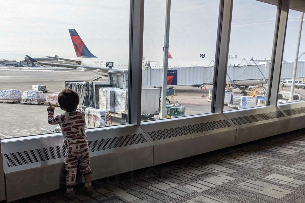 The image shows a toddler in fire truck-themed pajamas standing at an airport window, pointing at airplanes on the tarmac. The child is facing away from the camera, and outside the window, a Delta Airlines plane is parked at a gate, with cargo and equipment visible nearby. The scene captures the child's fascination with the planes.