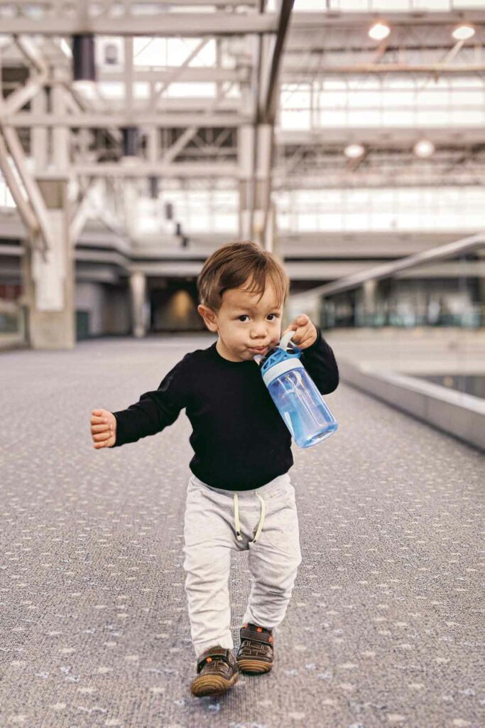 The image shows a toddler walking confidently through Denver airport’s mezzanine, holding and drinking from a blue water bottle. The child is dressed in a black long-sleeve shirt, light gray pants, and brown shoes, with the spacious, modern airport terminal creating a blurred backdrop. The child’s determined expression and purposeful stride capture a moment of independence during travel.